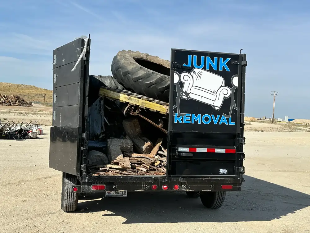 A black junk removal truck loaded with tires, wood, and debris. The back door displays a "JUNK REMOVAL" logo with a couch icon